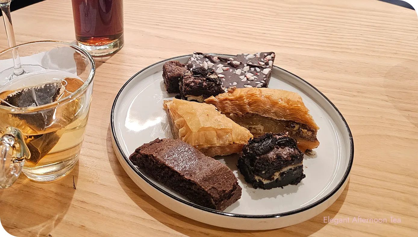 Dessert plate with baklava-style pastries, brownie bites, and chocolate bark beside tea and an iced drink