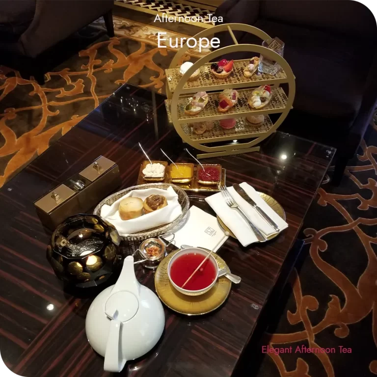 European afternoon tea served on a dark wood table with a gold tiered stand, teapot, scones, and preserves.